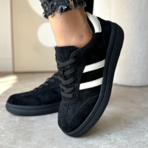 Czarne adidasy damskie LULA 97 wykonane z naturalnego zamszu, z białymi paskami i kontrastowym zapiętkiem, prezentowane na nodze w stylizacji z czarnymi jeansami.